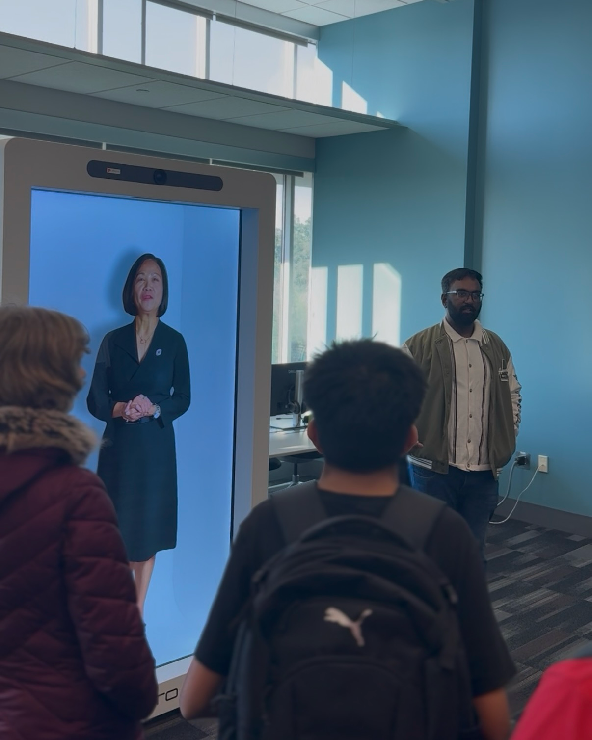 Omaha Central High School students stand in front of the Proto spatial display as UNO Chancellor Joanne Li appears at human scale, with Vishva Prasanth Srinivasan standing beside the display.